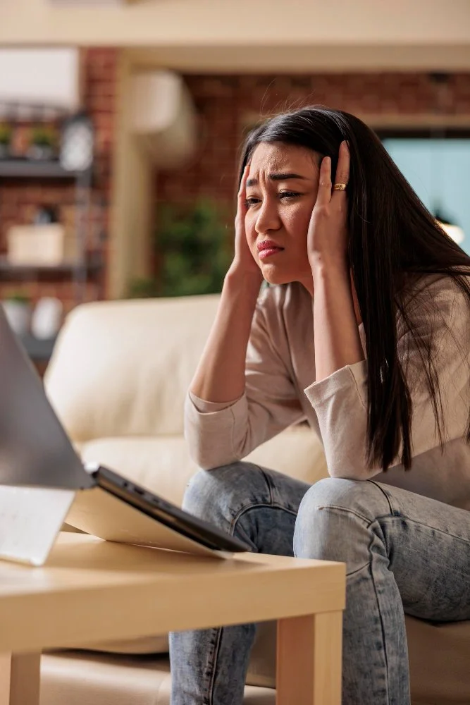 A woman sitting on a couch holding her head with both hands, looking distressed while looking at a laptop on a wooden coffee table.