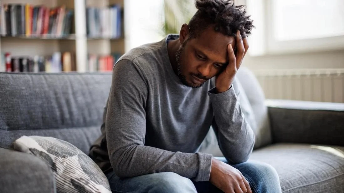 A man sitting on a gray couch with a distressed expression, hand on his head, in a room with a bookshelf in the background.