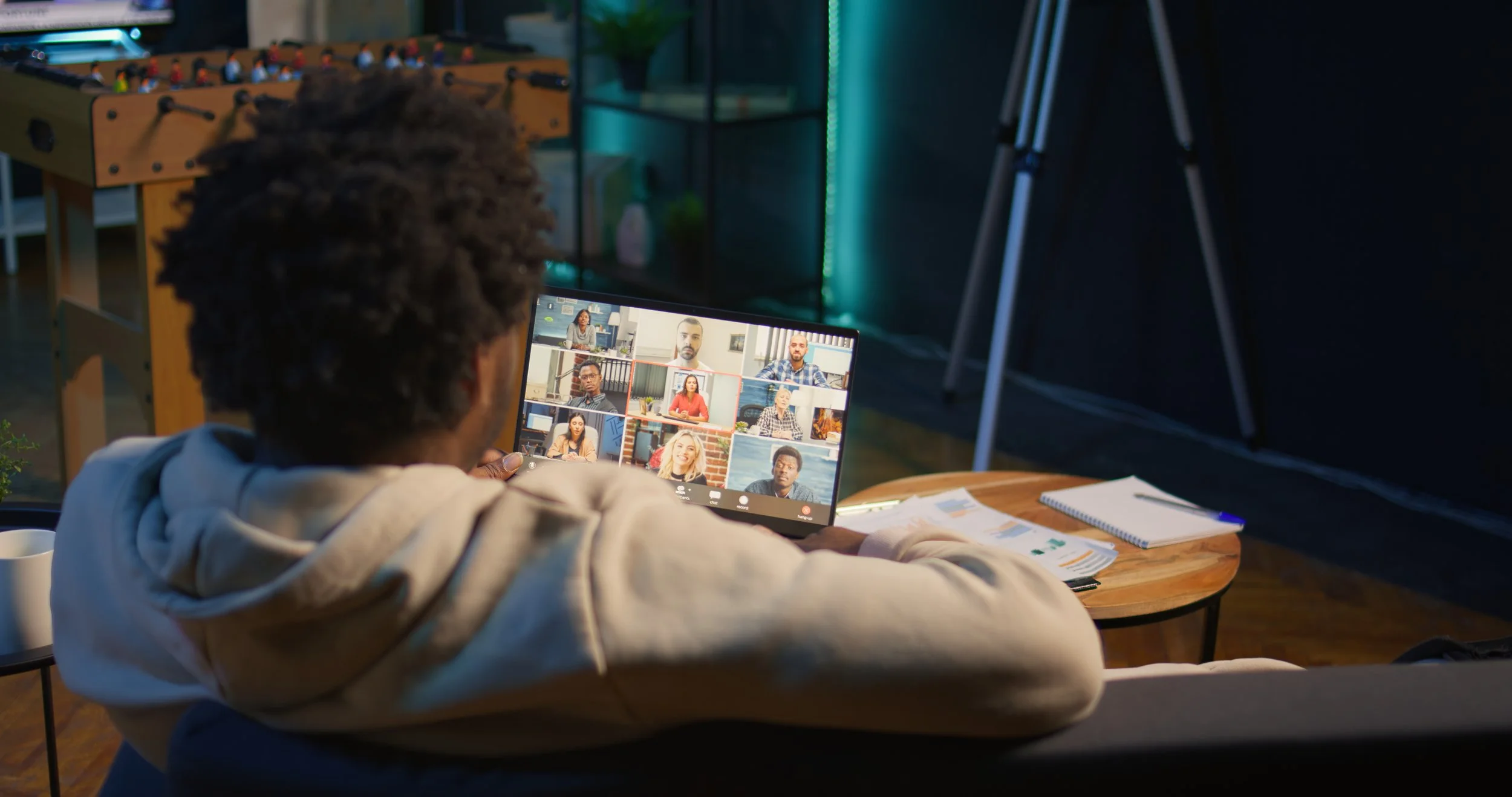 Person with curly hair participating in a virtual meeting on a laptop, with other meeting participants visible on the screen, in a professional indoor setting.
