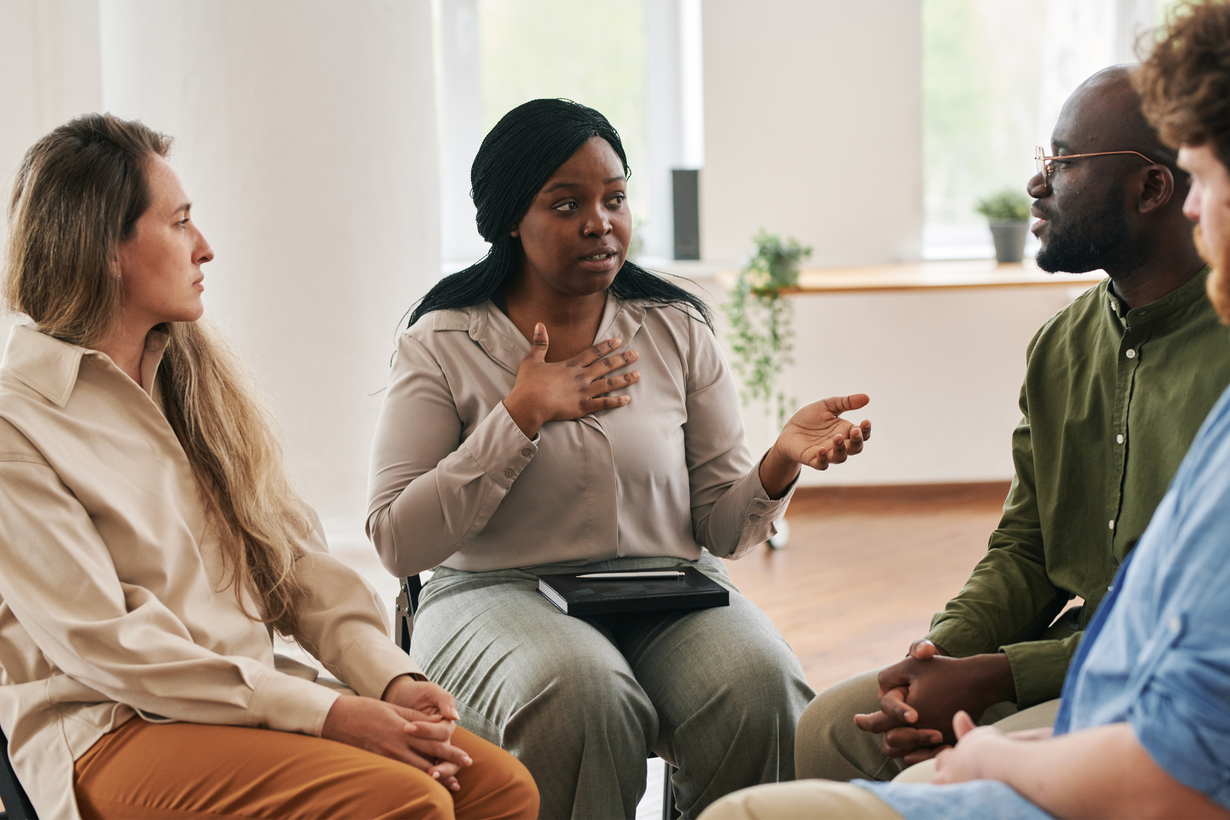 A diverse group of five people sitting in a circle, engaged in a discussion. The woman in the center is speaking, with her hand on her chest, and others are listening attentively.