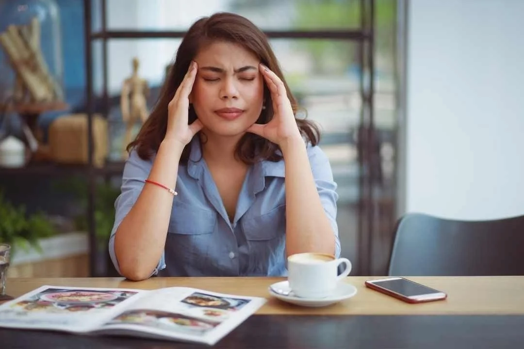 A woman sitting at a table with a magazine, a coffee cup, and a smartphone, holding her head with her eyes closed, appearing stressed or overwhelmed.