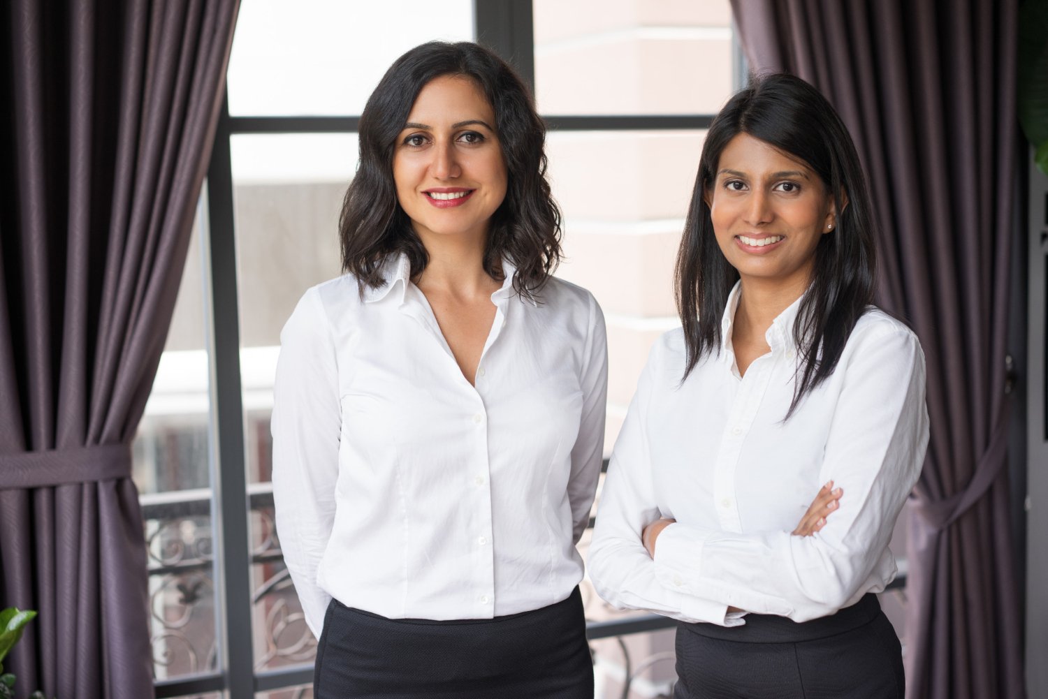 Two women in business attire standing indoors near windows with curtains, smiling at the camera.