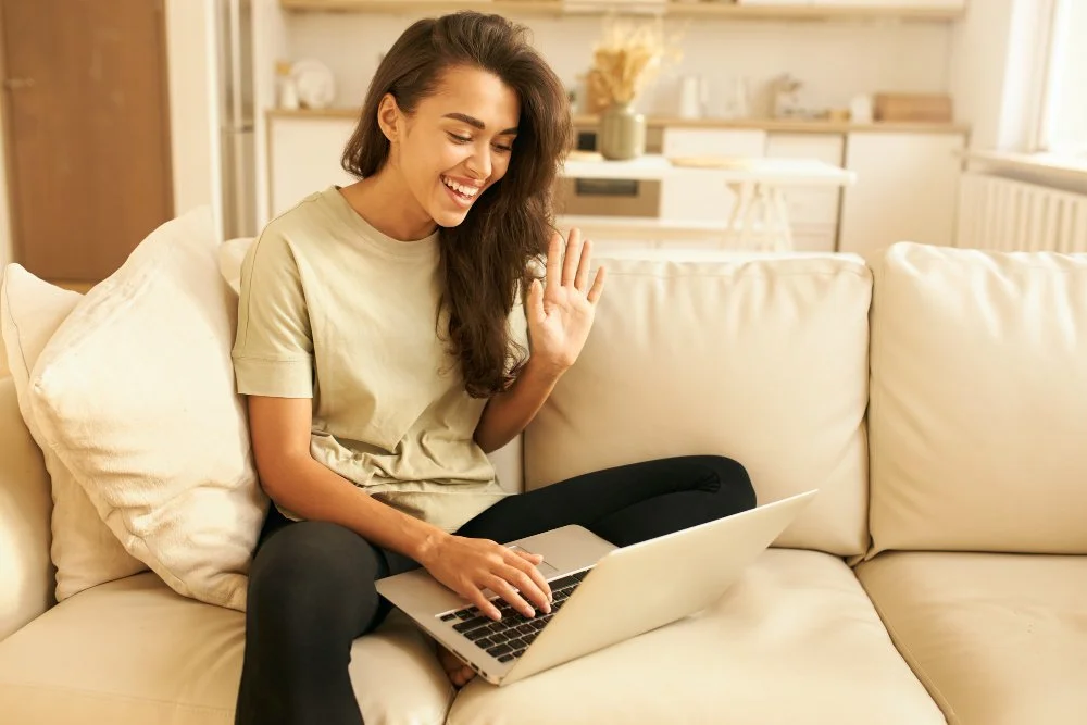 A woman sitting on a cream-colored sofa, smiling and waving at her laptop, in a bright living room.