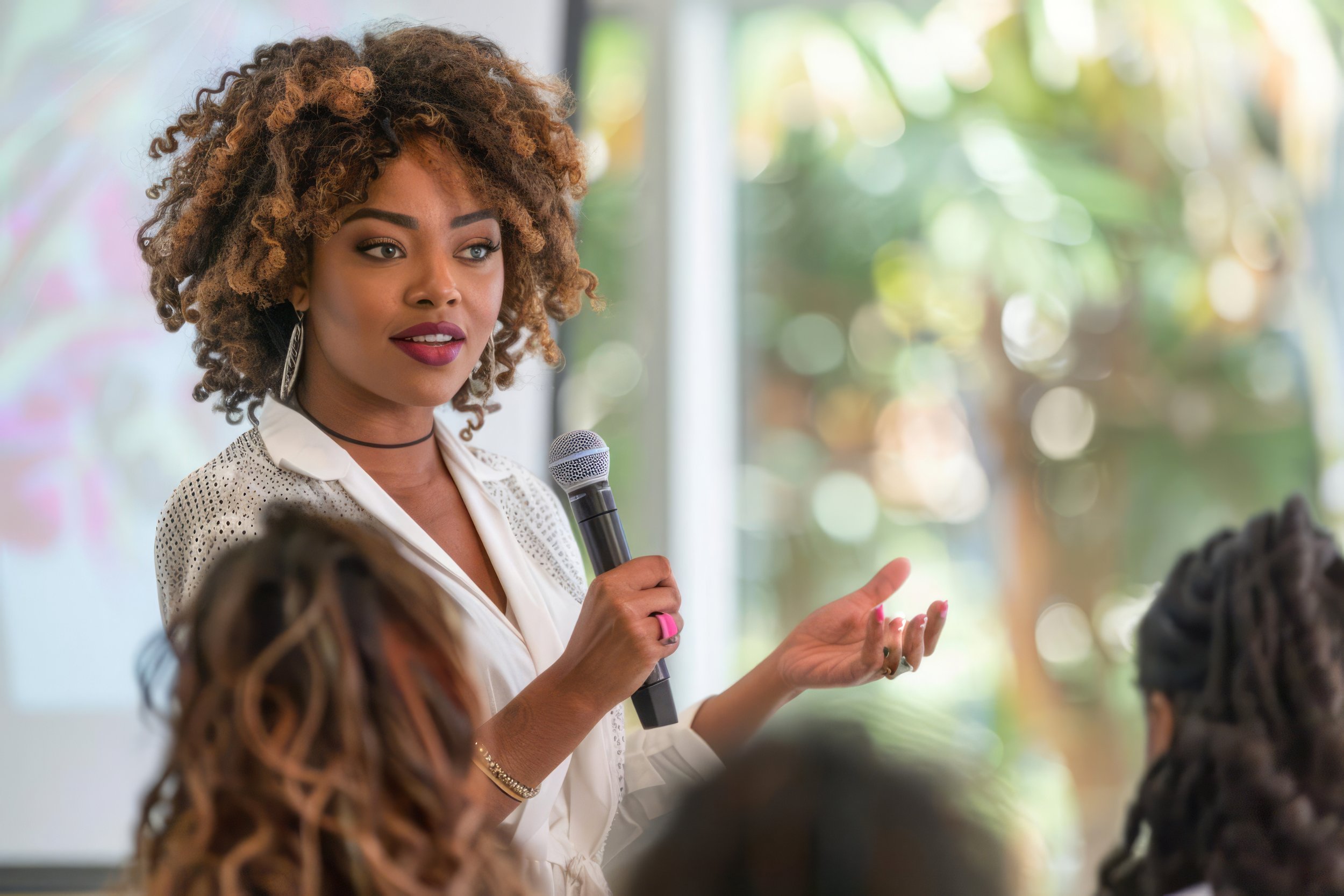 A woman with curly hair and makeup speaking into a microphone in front of an audience.