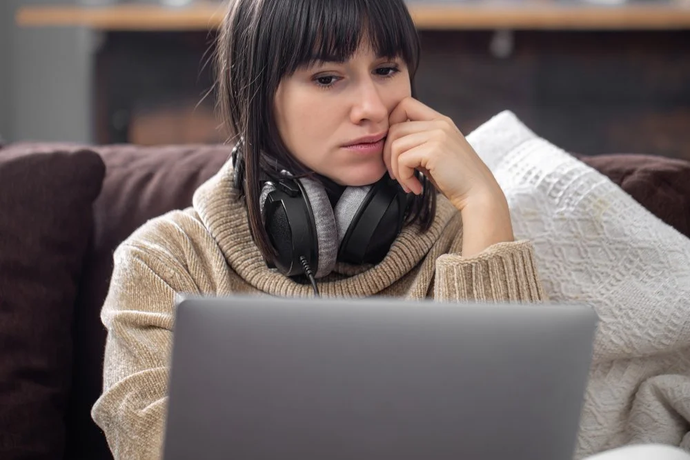 Woman with headphones sitting on a couch, looking at a laptop with a concerned or thoughtful expression.