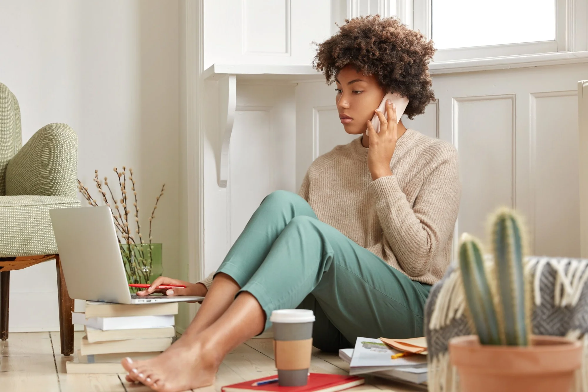 A woman with curly hair sitting on the floor by a window, talking on the phone, and working on her laptop among books and coffee cups.