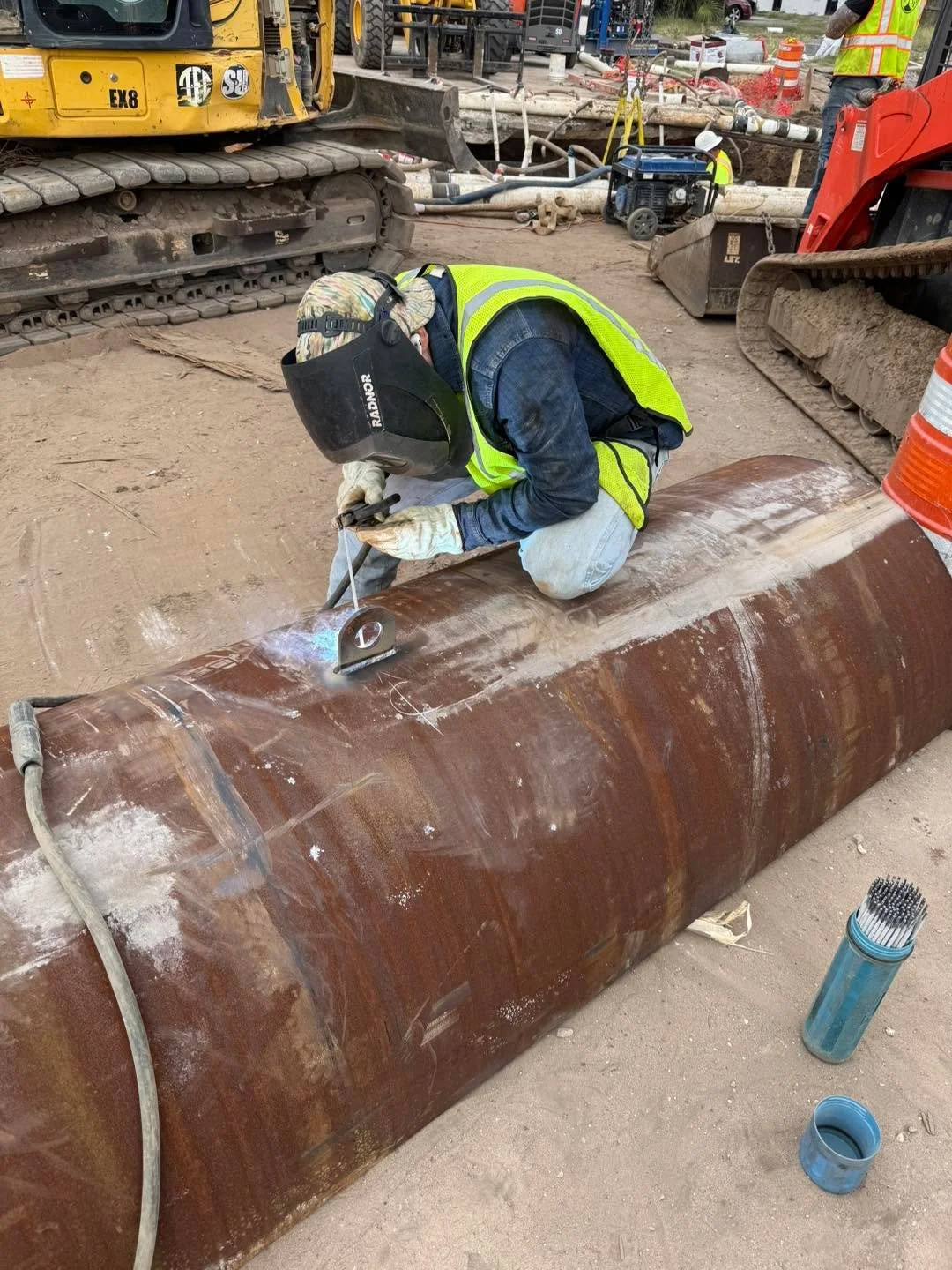 A worker welding a large, rusted metal pipe on a construction site. The worker is wearing a helmet, gloves, and a high-visibility safety vest, with construction machinery and workers visible in the background.