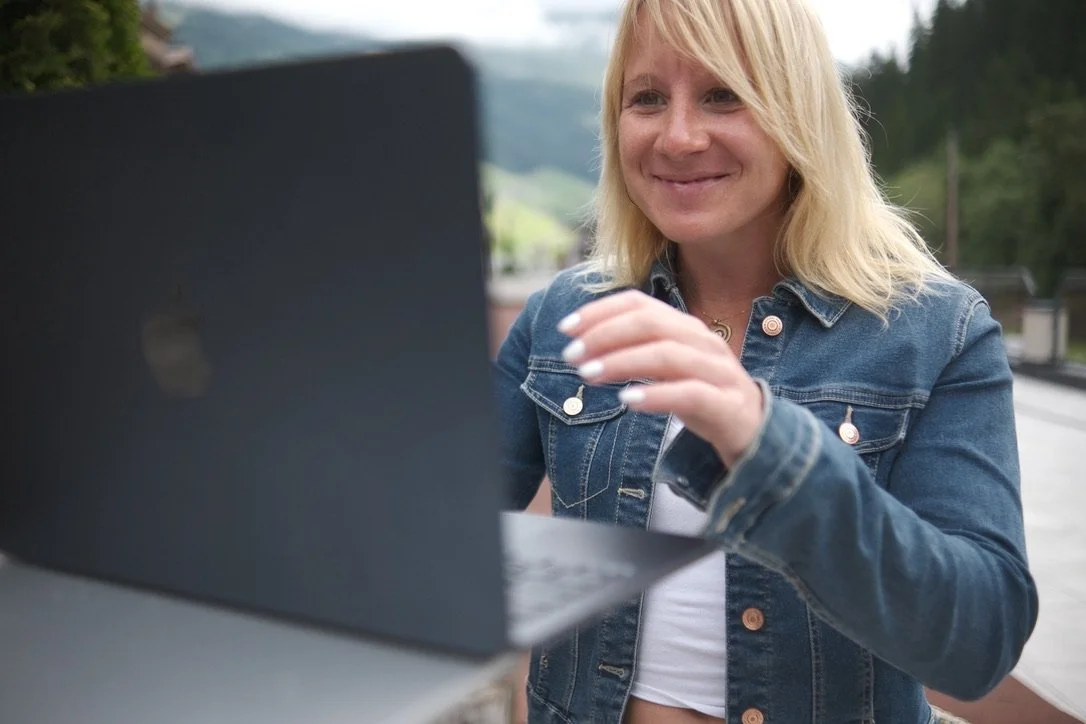 Frau lächelt, während sie draußen an einem Laptop arbeitet, mit einer grünen, hügeligen Landschaft im Hintergrund.