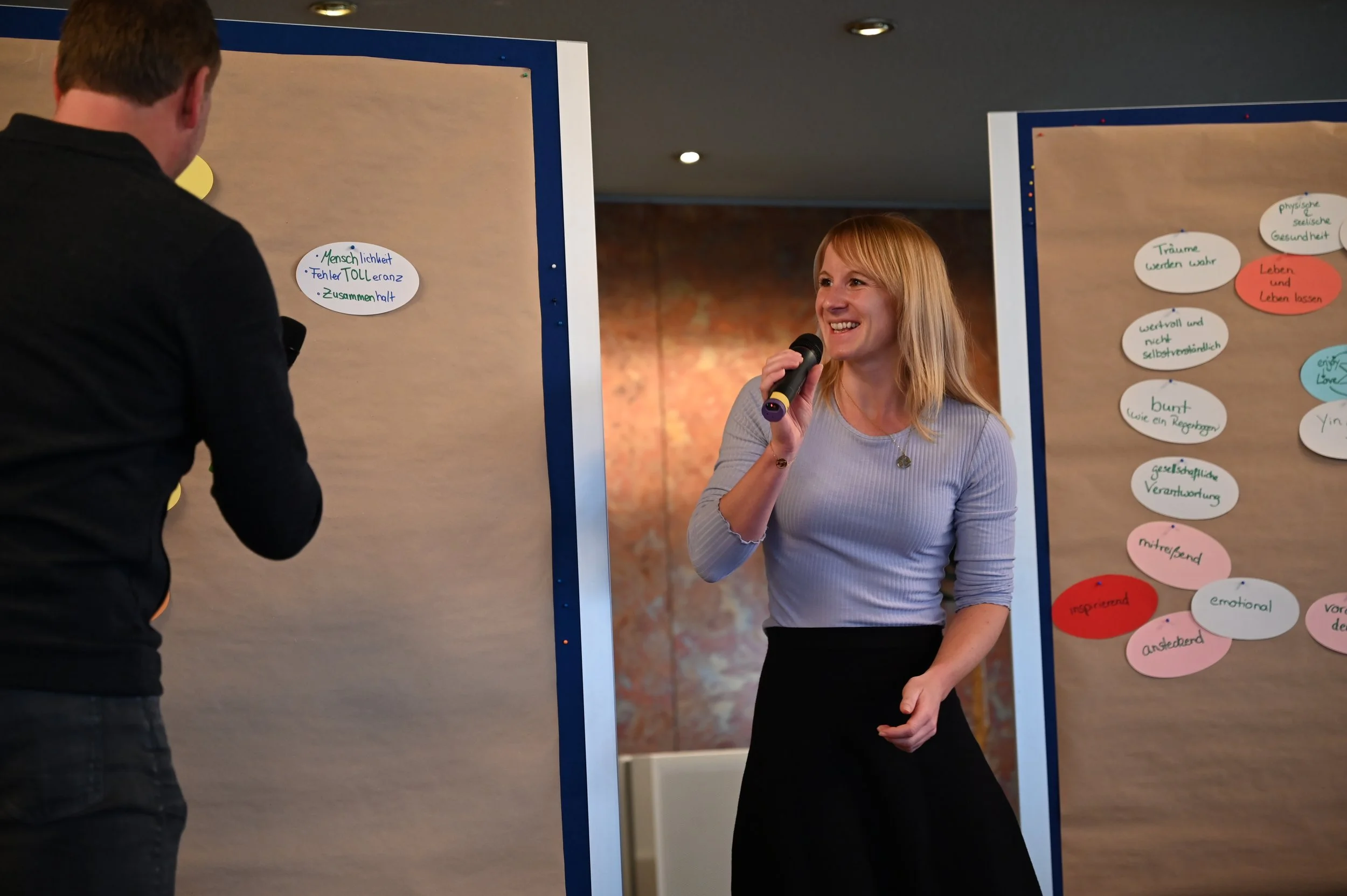 Eine blonde Frau hält ein Mikrofon und lächelt, steht vor zwei Pinnwänden mit bunten Notizen, während eine Person in einer schwarzen Jacke ihr gegenübersteht.