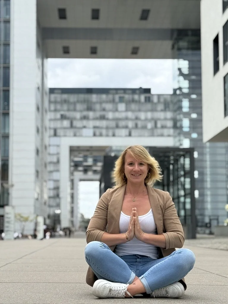 Eine Frau sitzt im Schneidersitz auf dem Boden in einer städtischen Umgebung, mit den Händen in Gebetshaltung, lächelt in die Kamera.