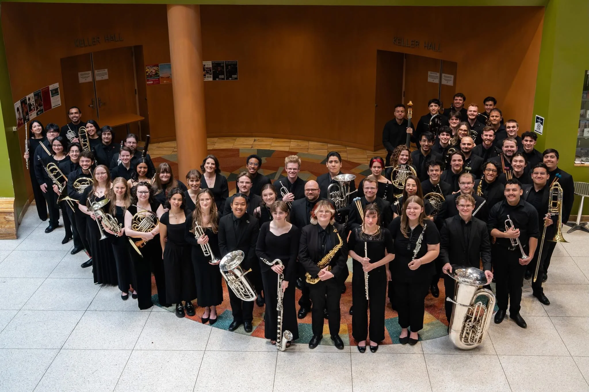 Large group of musicians holding various instruments, posing for a photo in an indoor hall with wood-paneled doors labeled 'Keller Hall'.