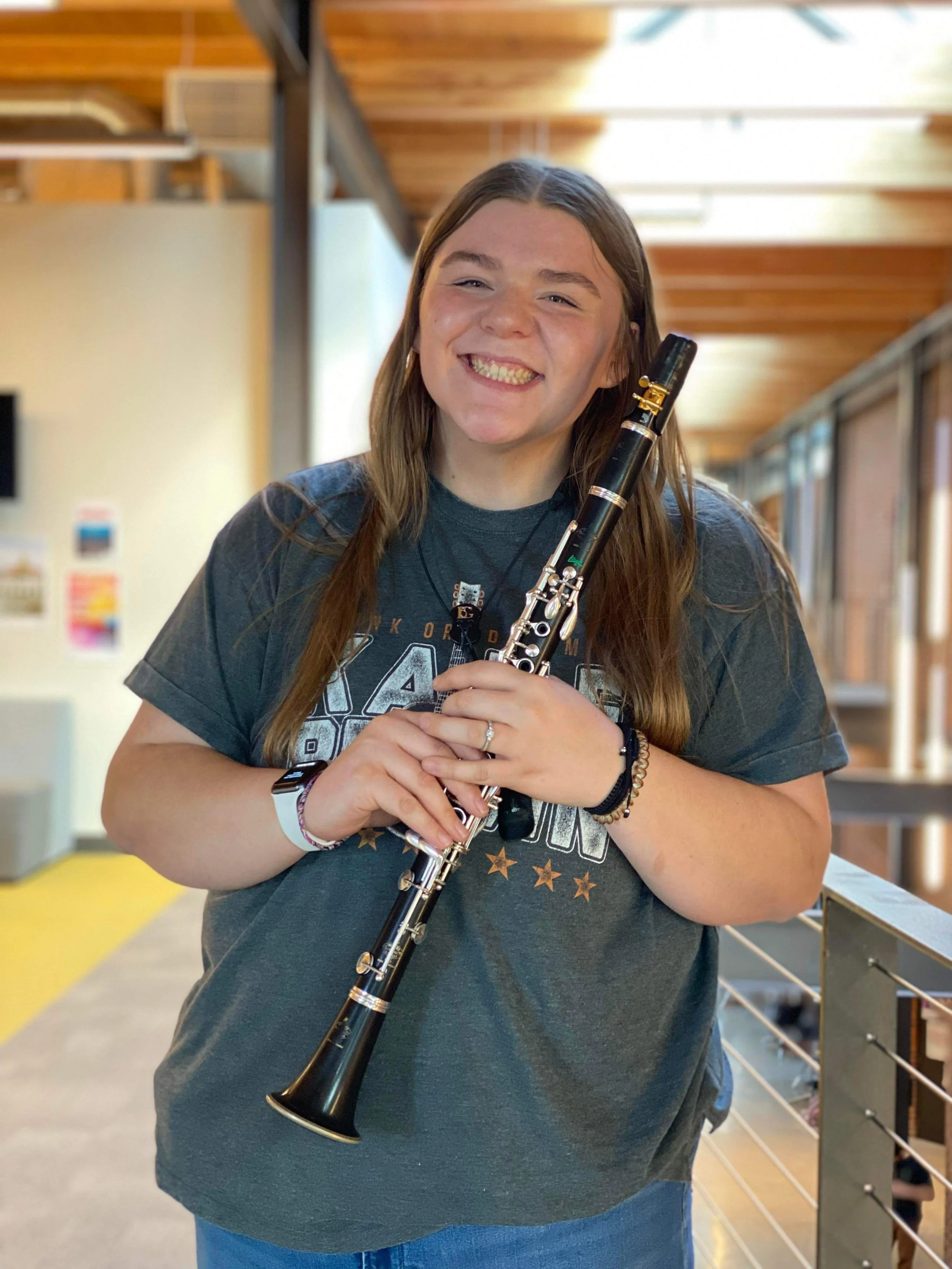 A young woman with long brown hair smiling while holding a clarinet in an indoor setting with wooden ceiling beams and beige walls.