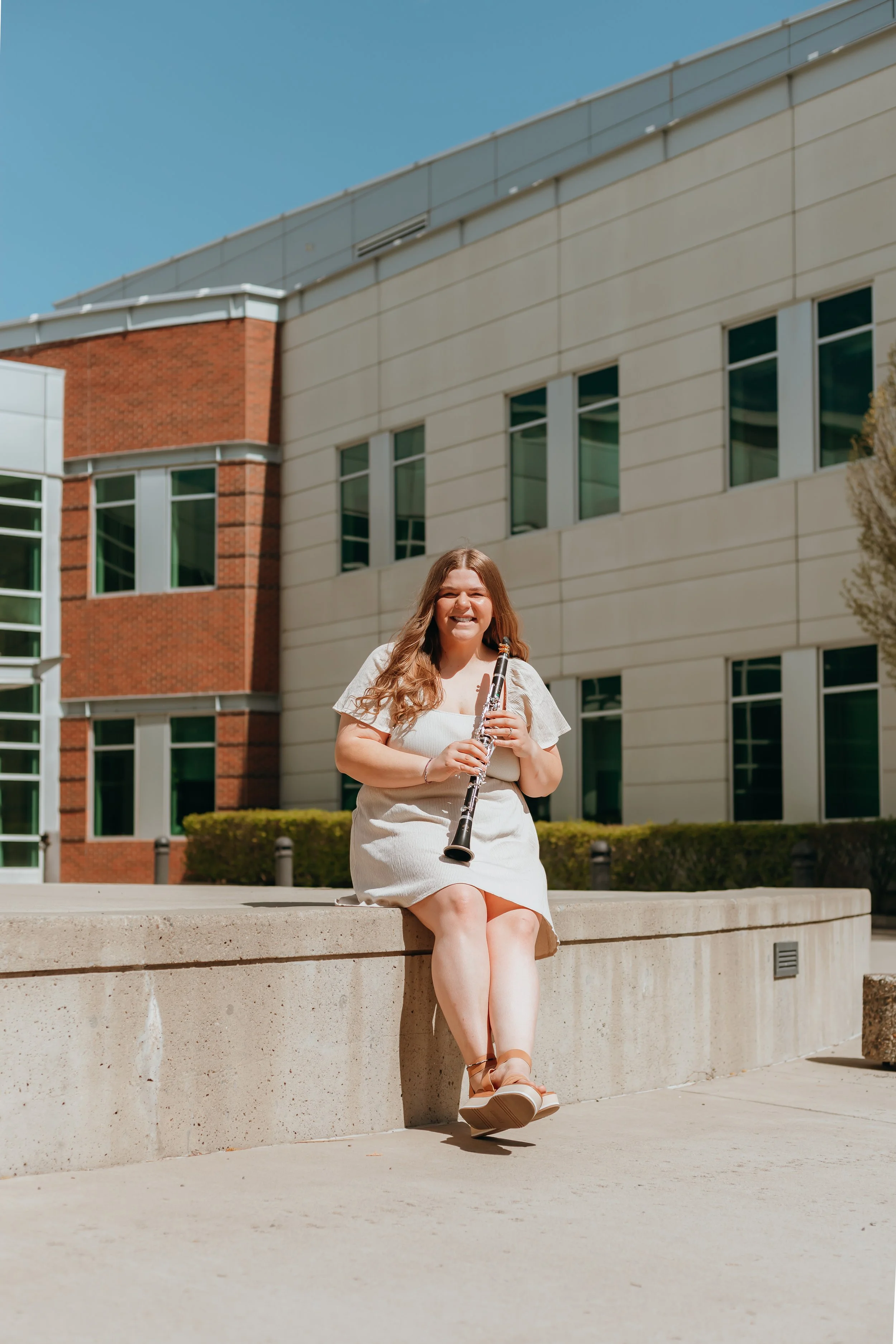 Young woman sitting on a concrete ledge, playing a clarinet outdoors in front of modern buildings, on a sunny day.