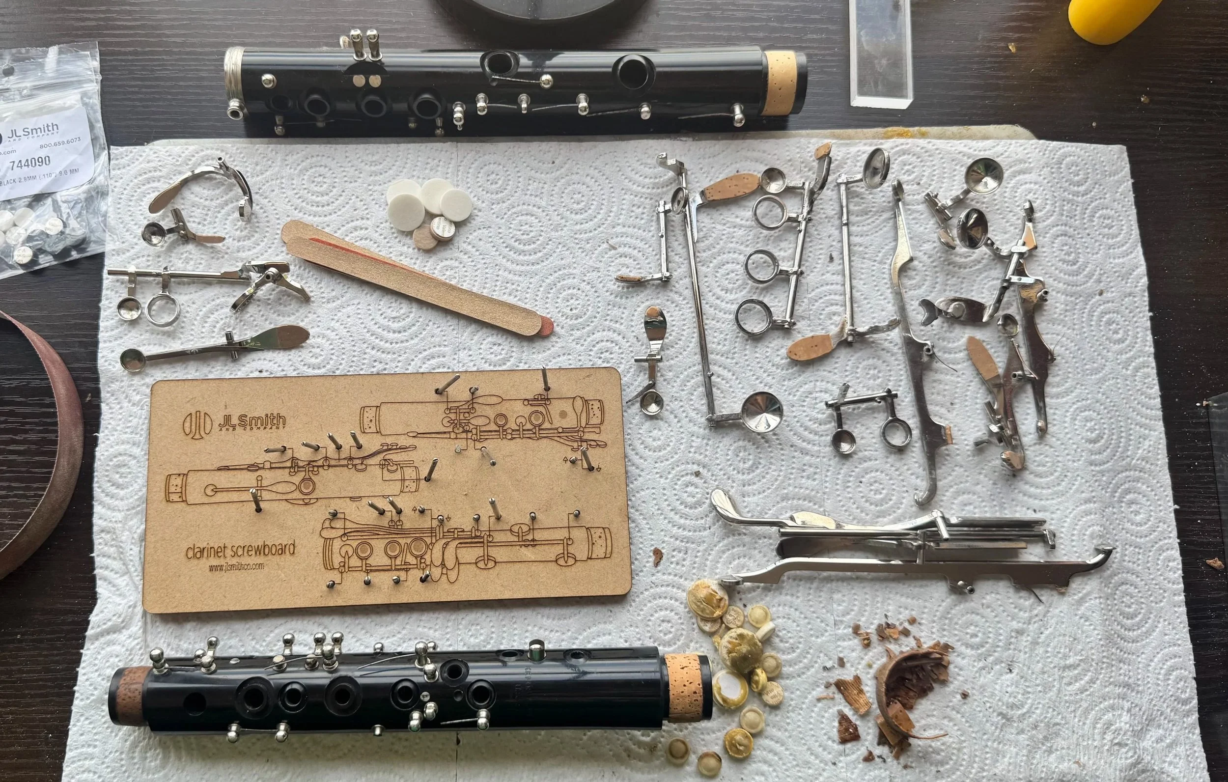 Disassembled clarinet parts and tools laid out on a paper towel, including two black hollow tubes, small metal components, pads, and a wooden clarinet diagram.