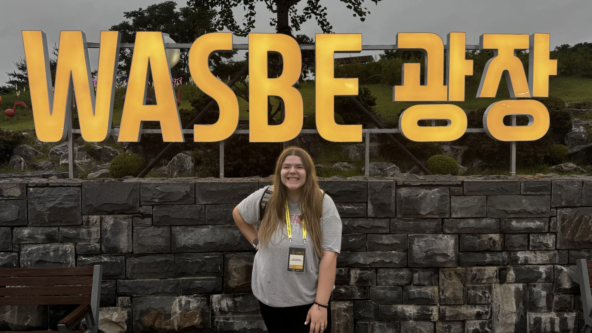 A young girl with long hair smiling and standing in front of a large yellow sign that reads 'WASDE' with reversed letters, on a stone wall with trees and cloudy sky in the background.