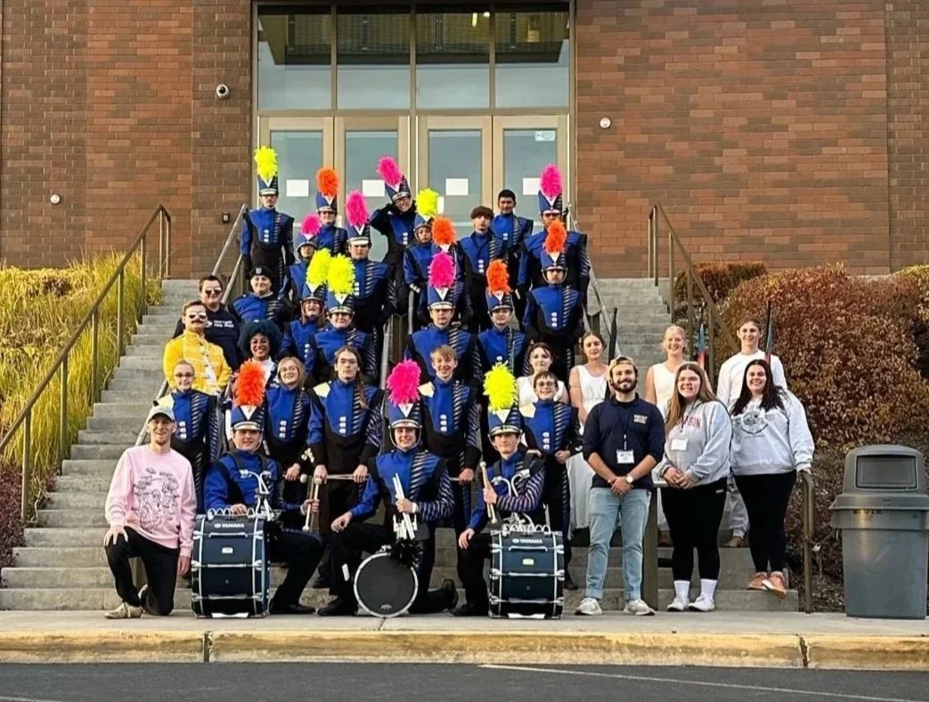 A marching band dressed in blue uniforms with colorful feathered hats stands on steps in front of a brick building, with some band members holding drums and musical instruments, and a few adults standing among them.