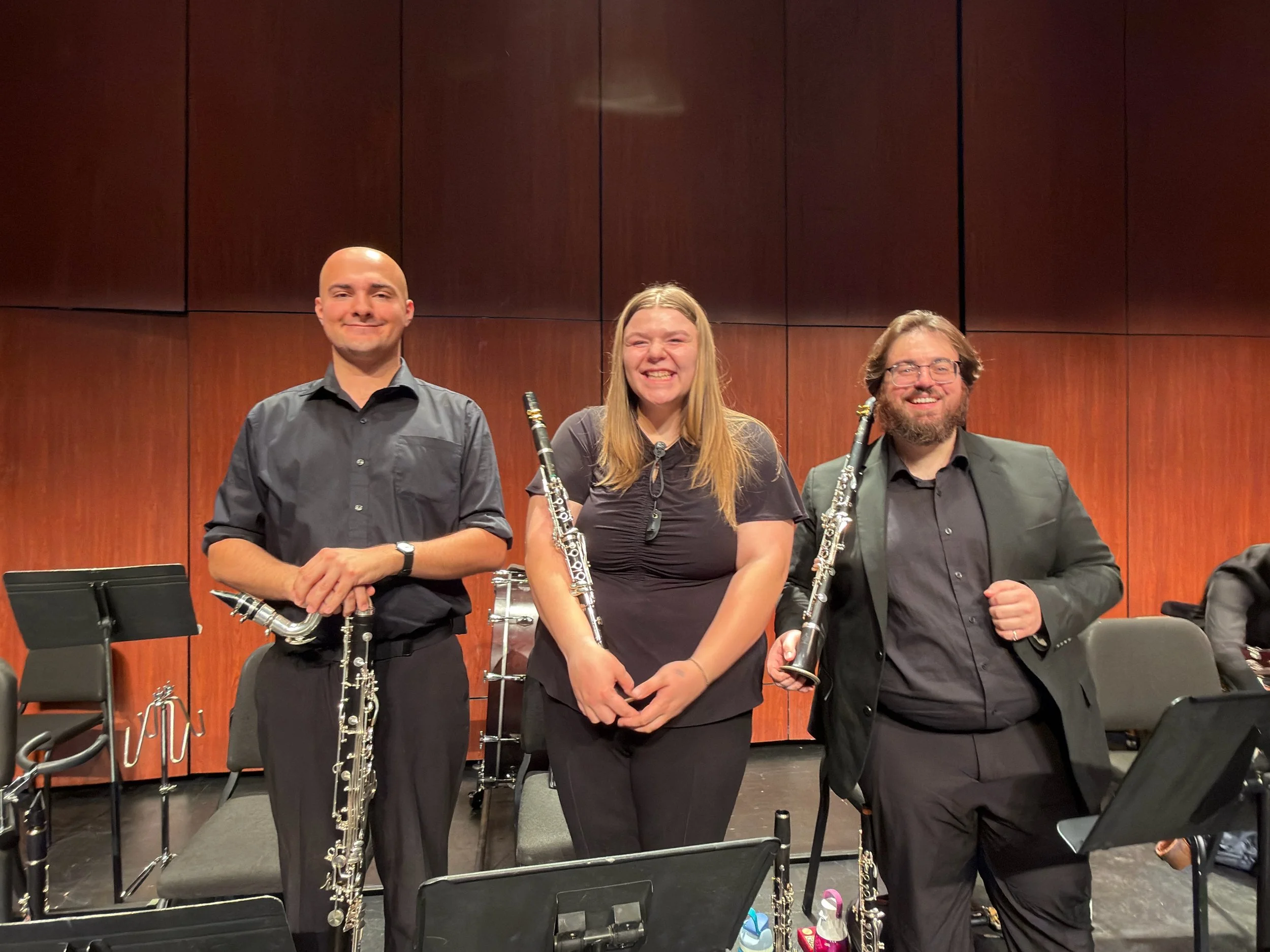 Three musicians standing on stage holding clarinets. They are in formal attire, with a wood-paneled backdrop and music stands in front of them.
