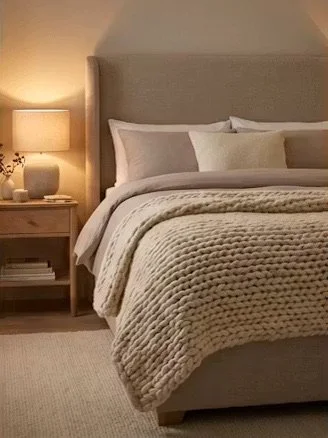 A neatly made bed with white pillows and beige bedding in a bedroom, beside a nightstand with a lamp and books.