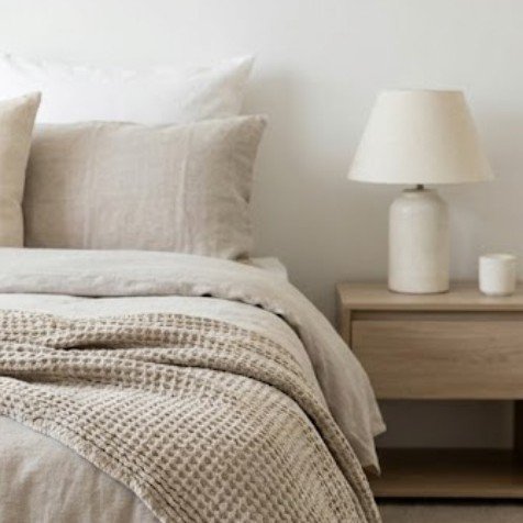 A bedroom corner with a neatly made bed, beige pillows, a textured blanket, and a wooden nightstand with a white table lamp and small decorative item.