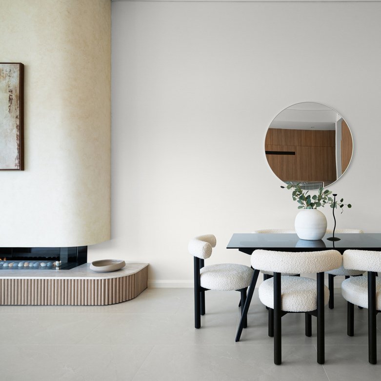 Modern dining room with a black table, four cream-colored upholstered chairs, a white vase with green foliage, a round mirror on the wall, and a contemporary fireplace with a curved beige wall.