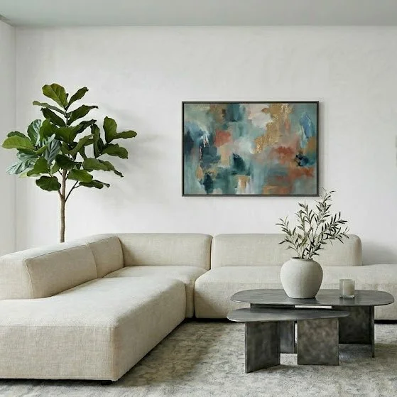 Modern living room with a large beige sectional sofa, a round black coffee table with a white vase and olive branch, a potted plant on a stand, a framed abstract painting on the white wall, and a light-colored carpet.