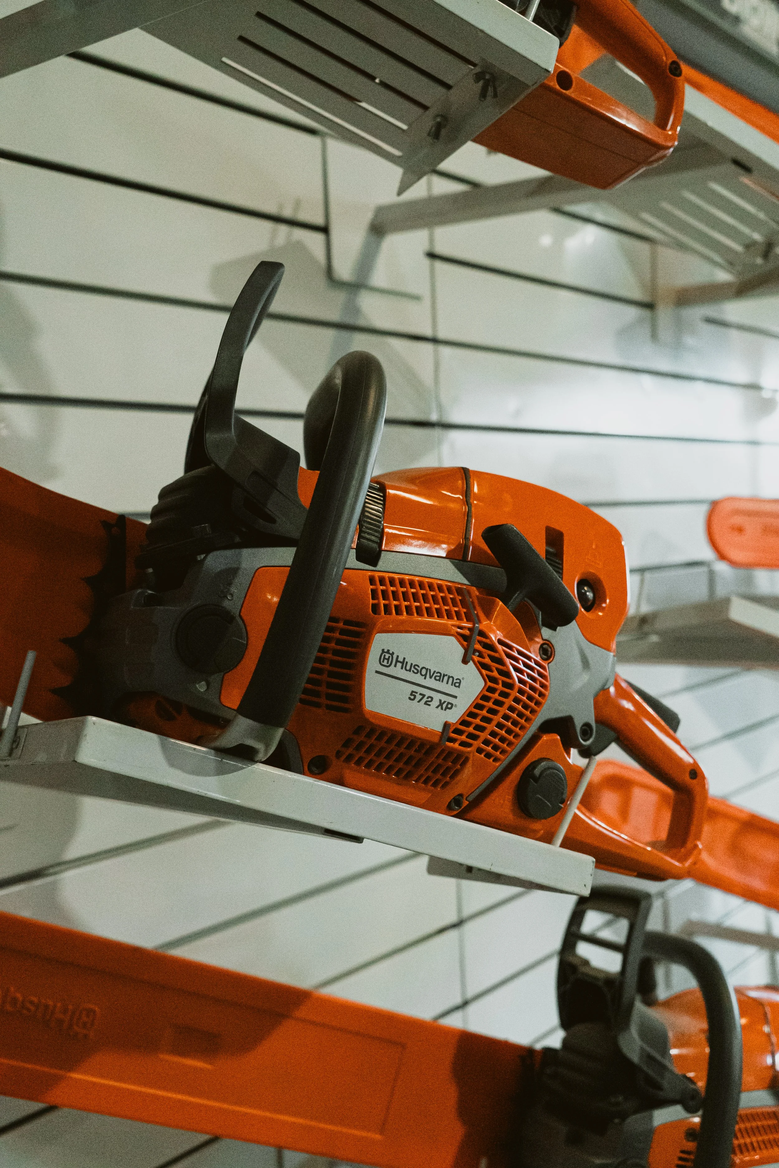 A chainsaw displayed on a white shelf. The chainsaw is orange with black controls, and it has a label that reads 'Husqvarna 572 XP'.
