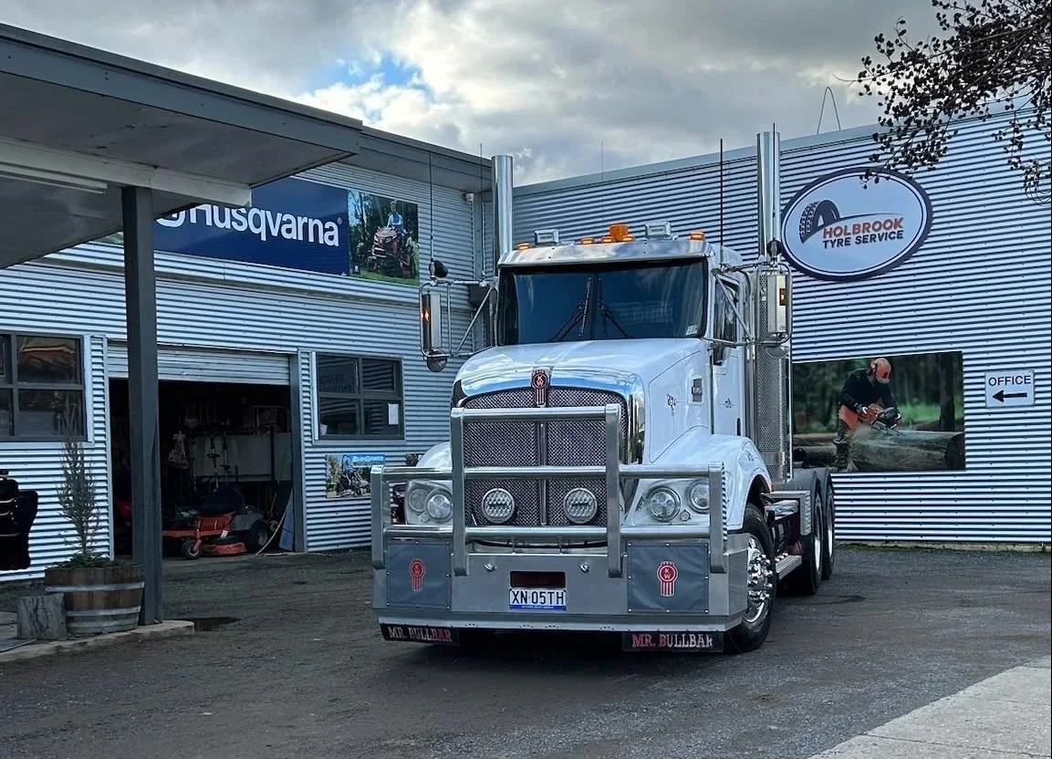 A large white semi-truck parked outside a building with signs for Husqvarna and Holbrook Tyre Service.