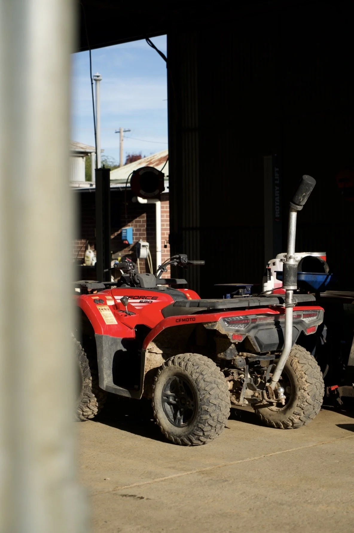 Red ATV parked outside a barn with tools and equipment visible inside. The background shows a blue sky and nearby rooftops.