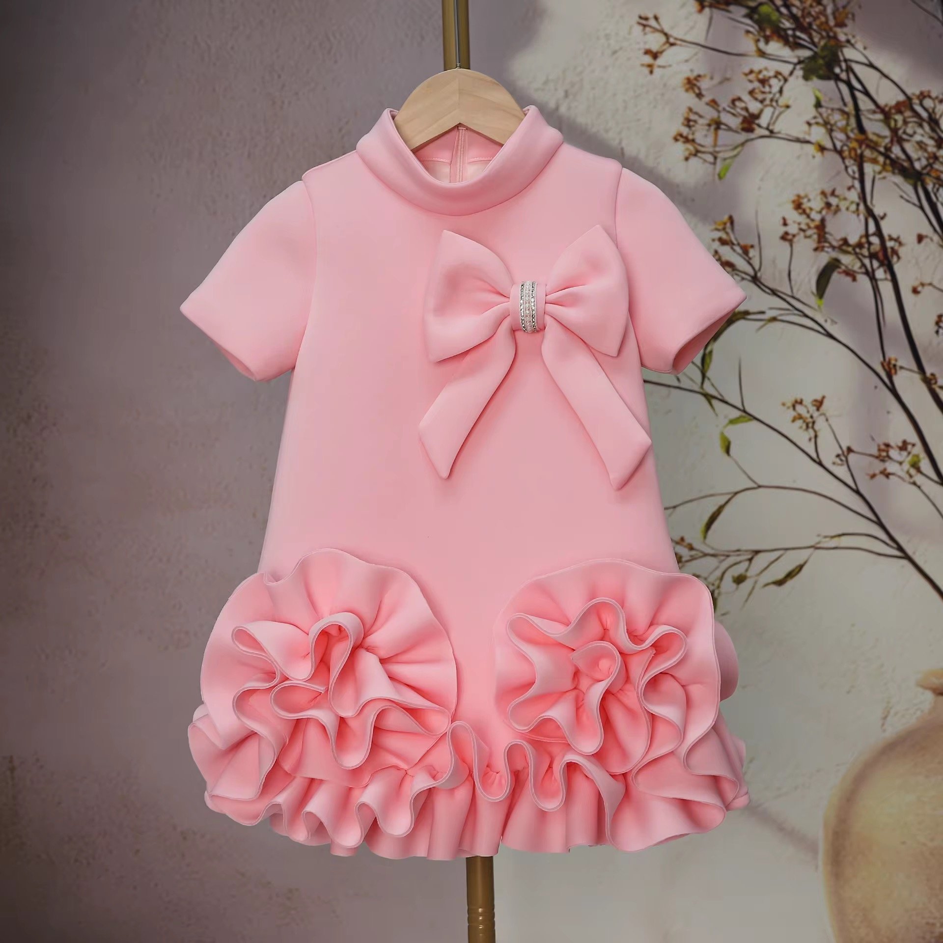 A pink dress with a large bow and rhinestone embellishment displayed on a wooden hanger against a wall with a floral mural.