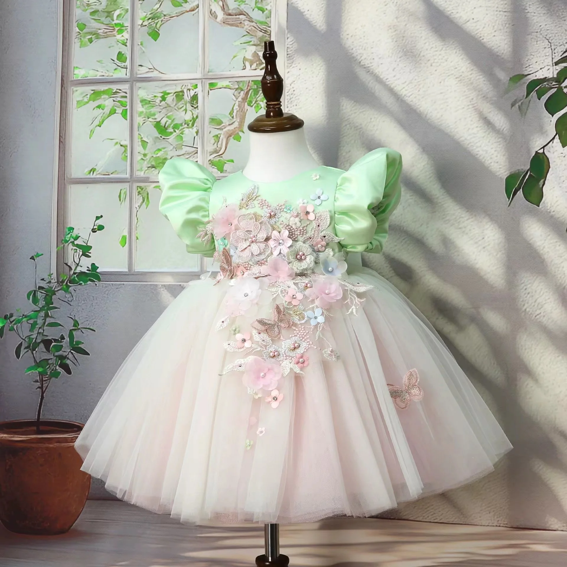 A dress displayed on a mannequin with puffed green sleeves and floral embroidery, set against a background featuring a window, a potted plant, and a textured wall.
