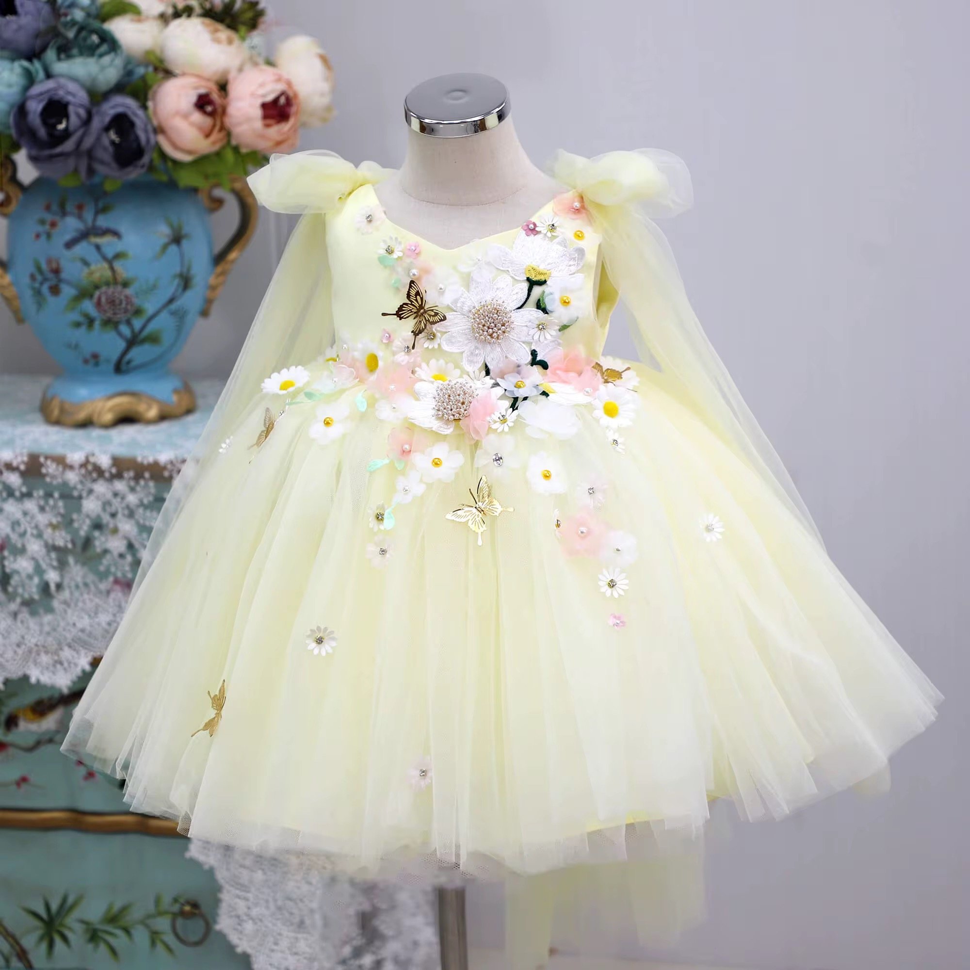 A white mannequin dressed in a pale yellow dress with floral and butterfly embellishments, including lace and pearl details, displayed in a boutique or showroom. To the left, a decorative vase with a floral pattern holds a bouquet of pastel-colored flowers.