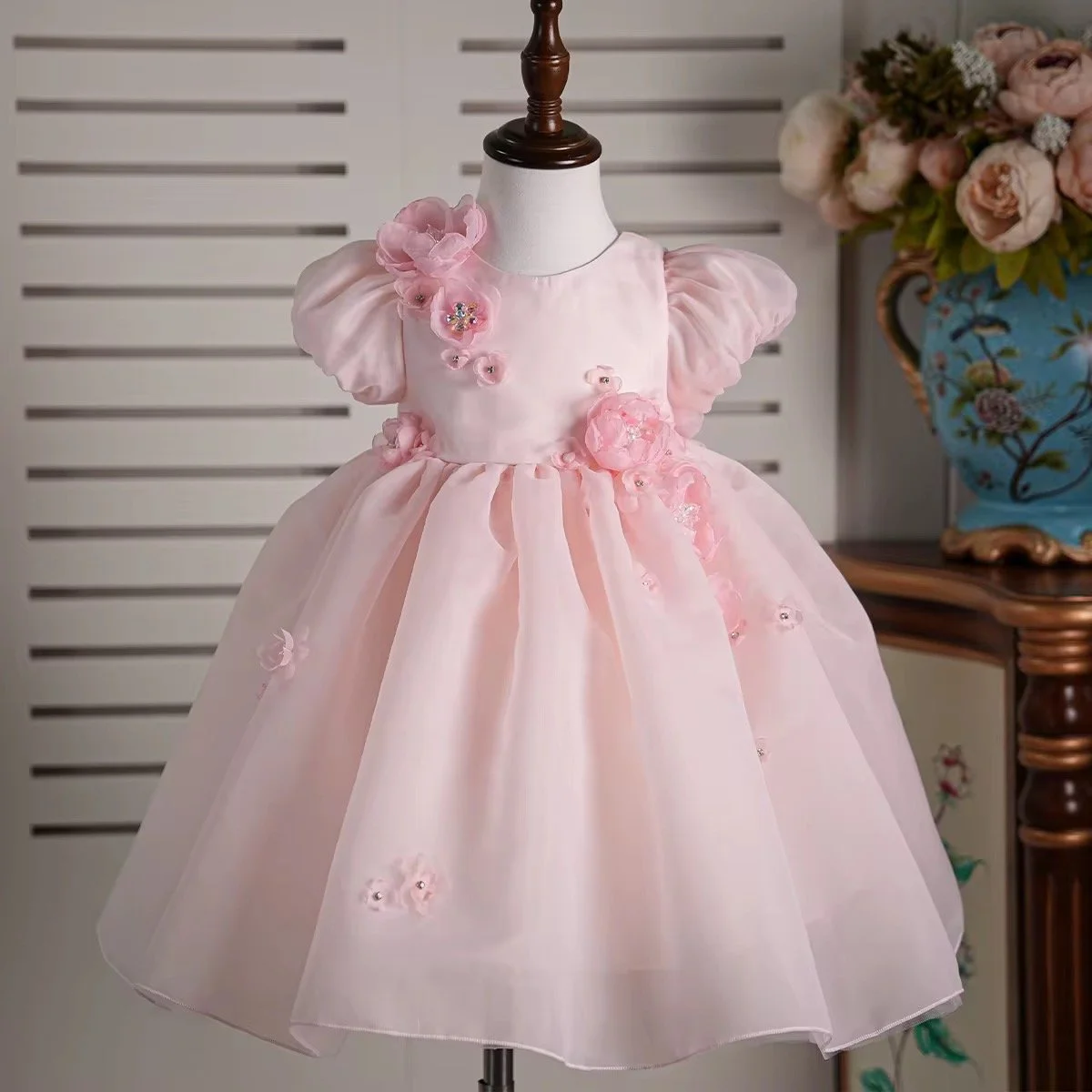 A pink dress for a young girl, displayed on a white mannequin with a dark wood top. The dress features puffed short sleeves, floral appliqués, and a full skirt made of light pink fabric.