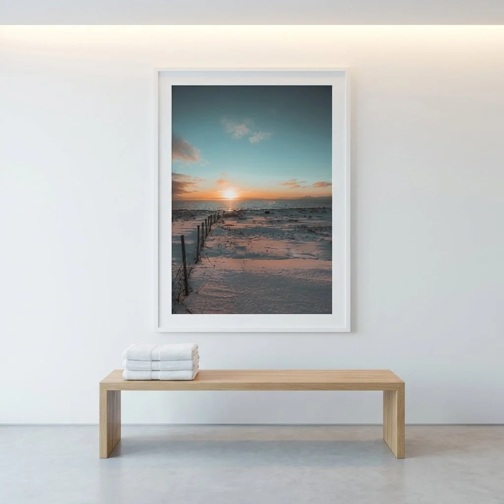 A framed picture of a sunset over a sandy beach hangs on a white wall in a minimalist room, with a wooden bench and folded towels beneath it.