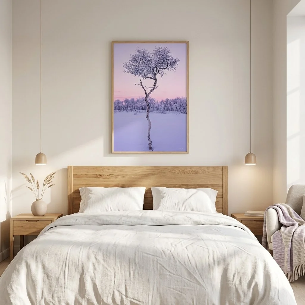 A bedroom with a wooden bed frame, white bedding, two matching pillows, and two small hanging pendant lights. A framed picture of a bare tree in a snowy landscape with a pink and purple sky hangs above the bed. There are nightstands with vases and books, and a cozy armchair with a blanket on the right side.