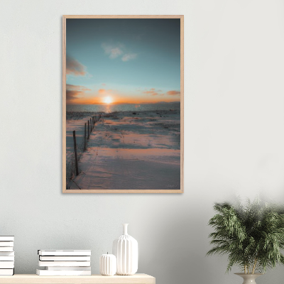 A framed photograph of a snow-covered beach at sunset, with a wooden fence partially buried in snow, hung on a white wall in a modern interior setting.
