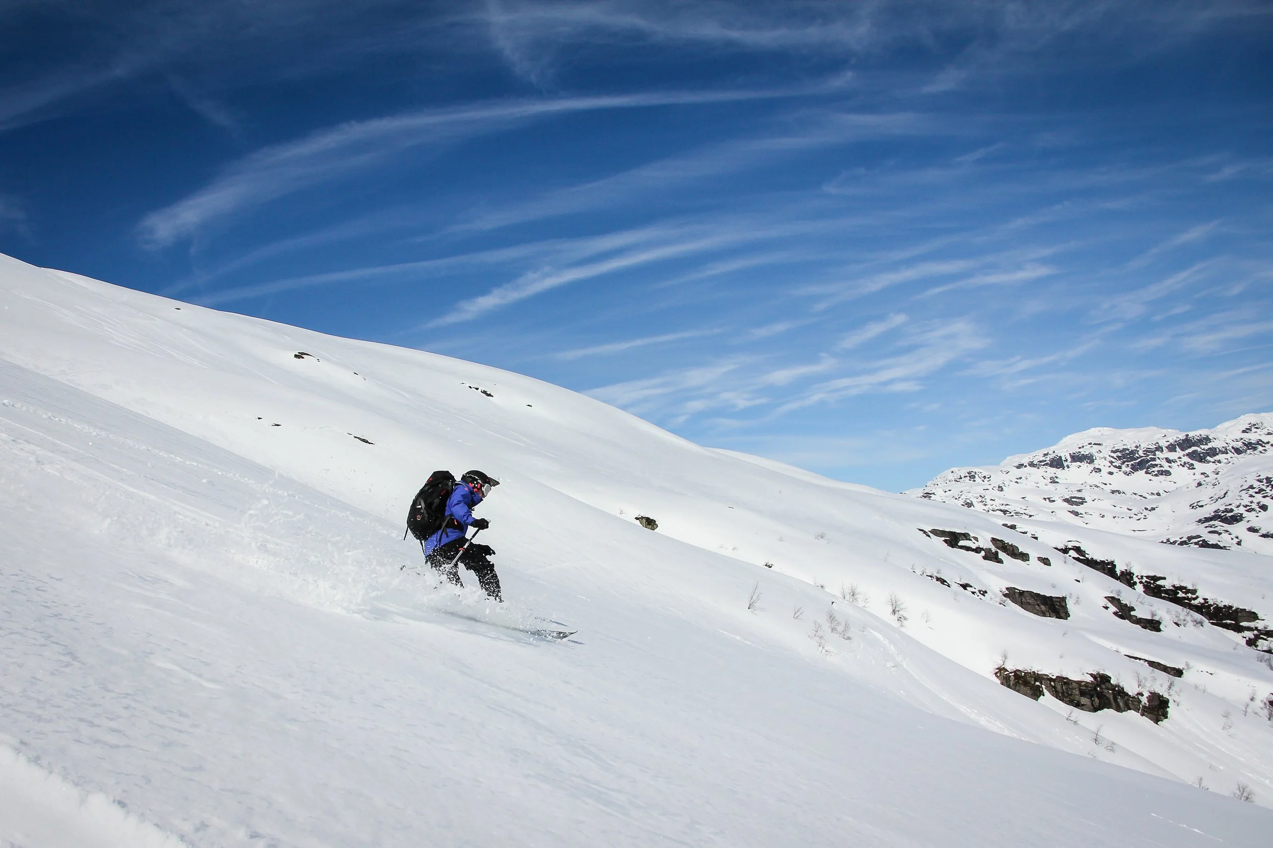 A person in a blue jacket skiing down a snowy mountain slope with a backpack, under a partly cloudy blue sky.
