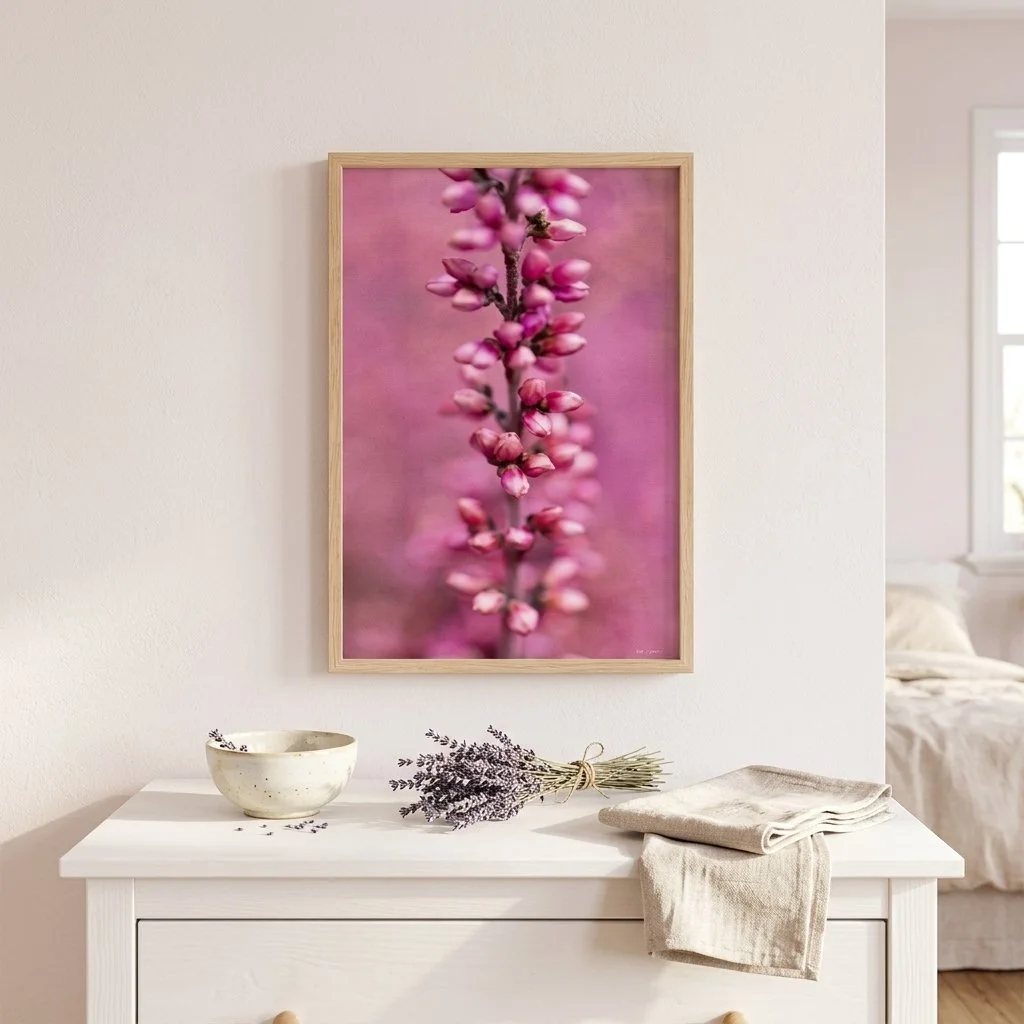 A framed pink floral art print hanging on a white wall above a white dresser with a bowl, lavender sprigs, and folded linens.