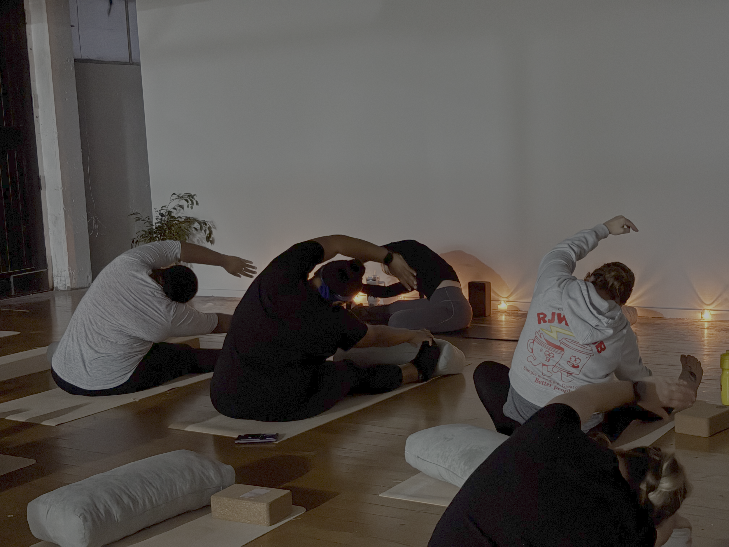A group of people practicing yoga or meditation in a dimly lit room with candles, lying on mats in a seated forward bend position, with a plain wall and potted plant in the background.
