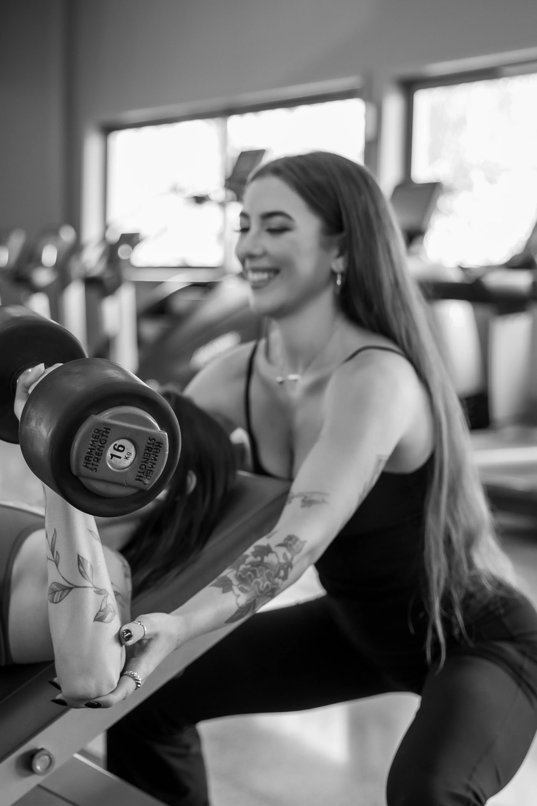 A woman with long hair, tattoos, and earrings is in a gym, lifting a dumbbell with her right arm while smiling with her eyes closed. The gym has large windows in the background.