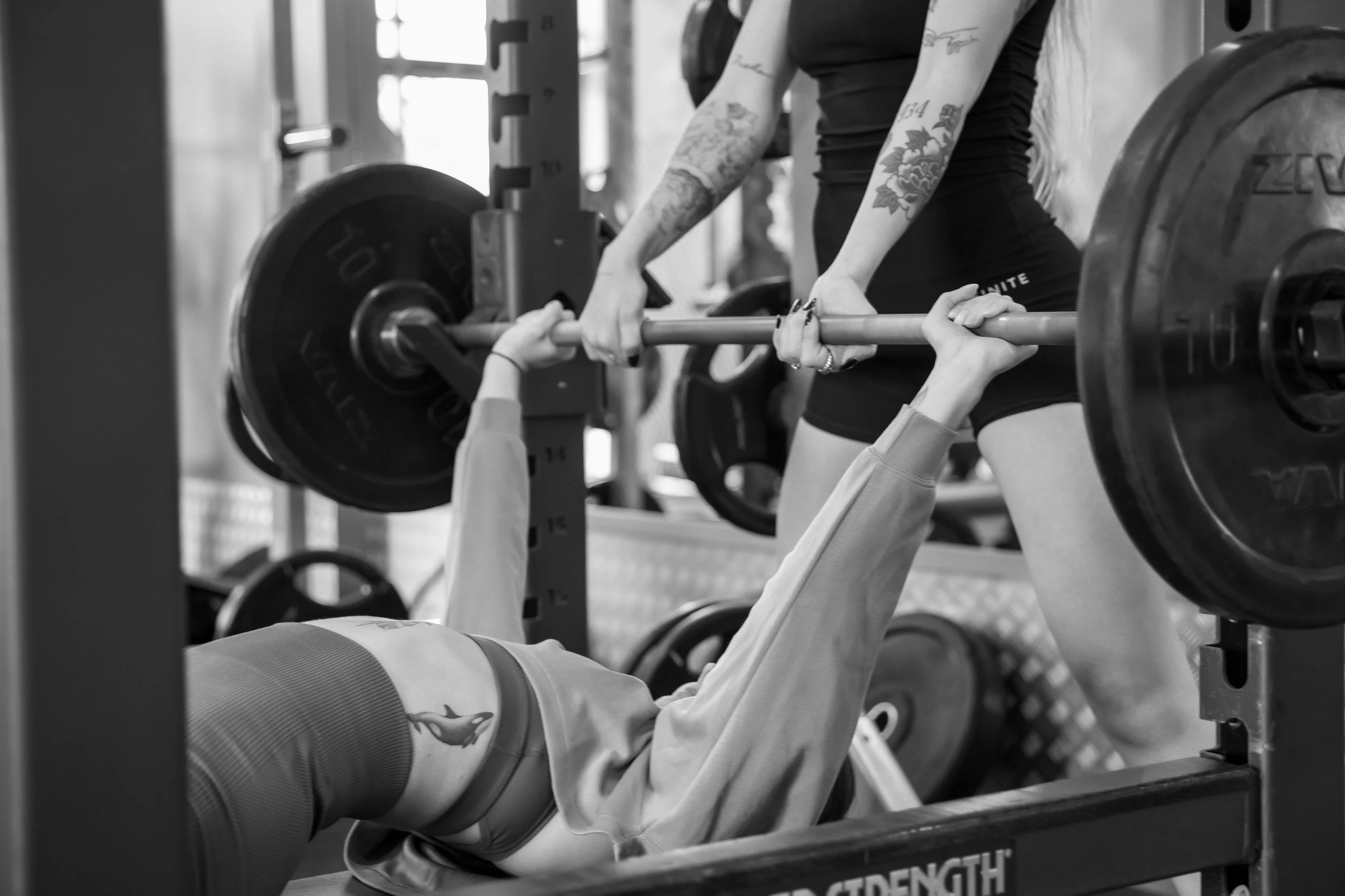 A person with tattoos on their arms lying on a bench press at a gym, lifting a barbell with weights.