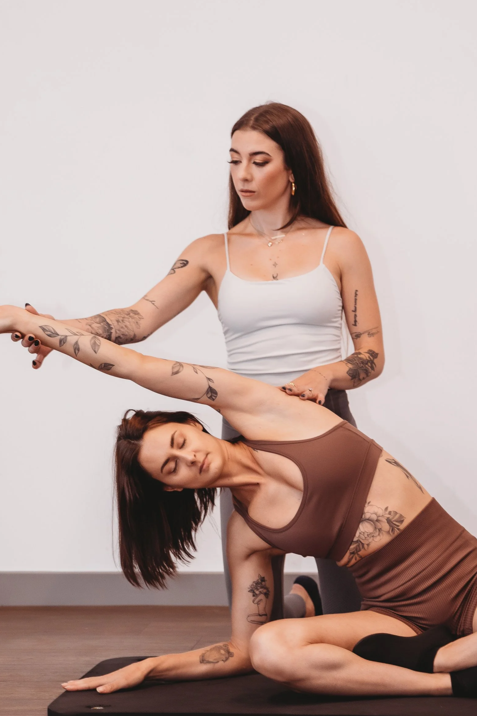 Two women practicing yoga together indoors, one standing and the other lying on a yoga mat, both in a yoga pose. They have tattoos and are focused on their practice.