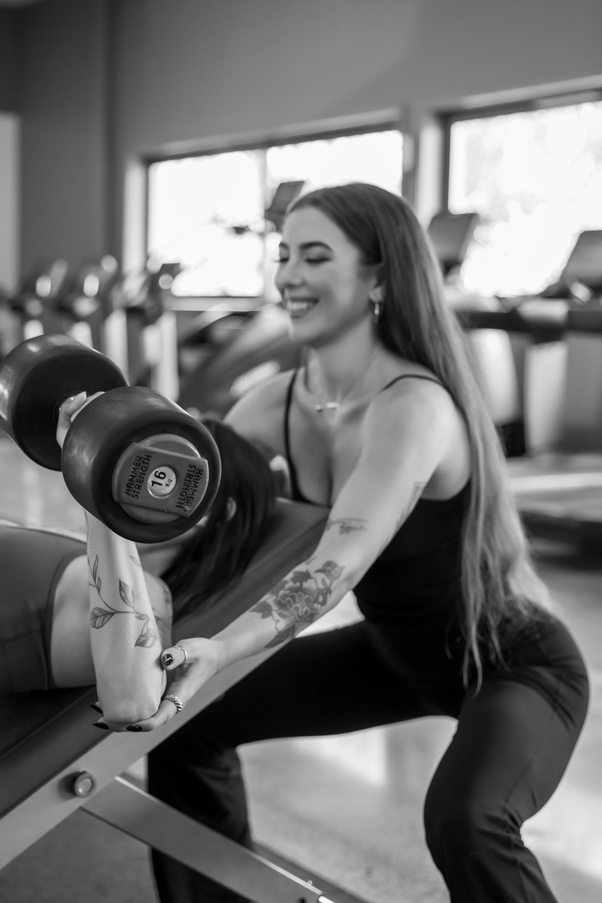 A woman with long hair, tattoos on her arms, and wearing a black tank top is sitting on a workout bench, lifting a dumbbell in a gym.