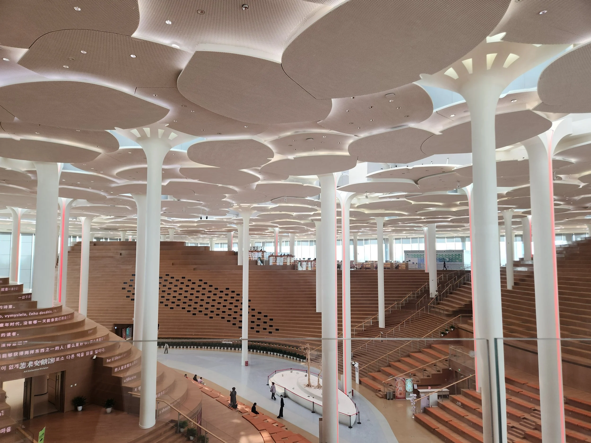 Interior of a modern building with a high ceiling featuring large, cloud-shaped white structures supported by white columns. Wooden stairs and platforms are visible, along with some people walking around and sitting.