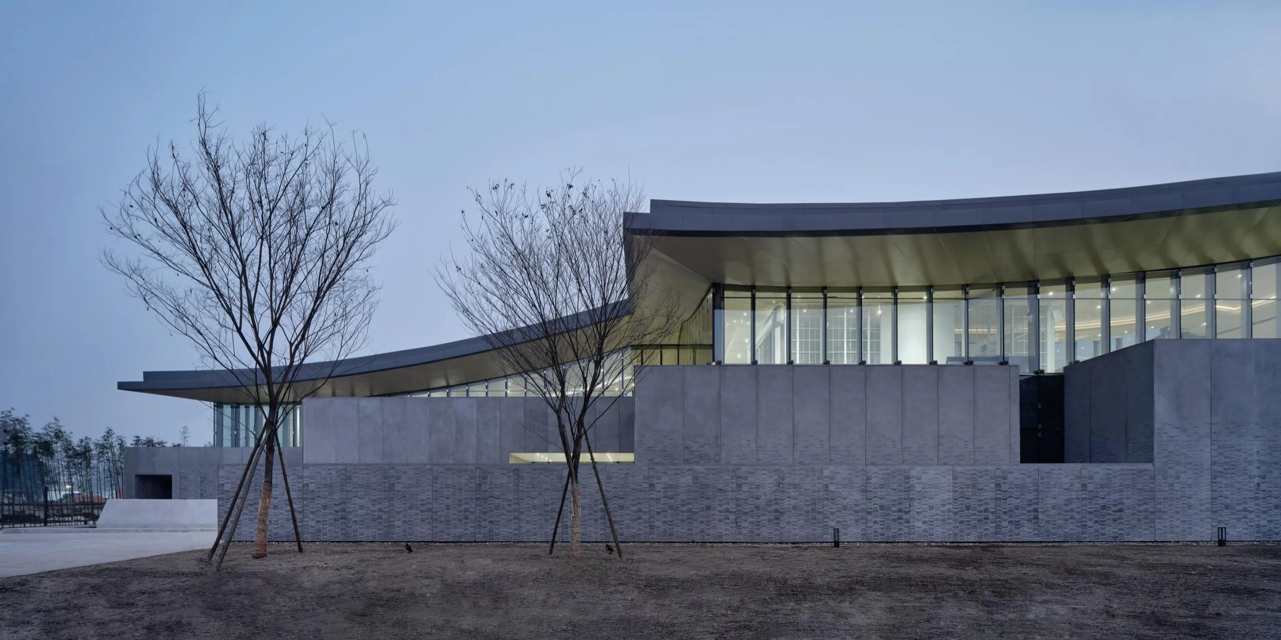 Modern building with large glass windows and a wave-shaped roof, surrounded by leafless trees at dusk.