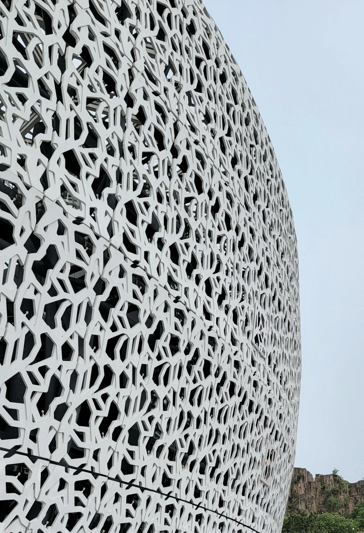 A modern building with a decorative façade made of interconnected white patterns, set against a clear sky and distant rocky hills with greenery.