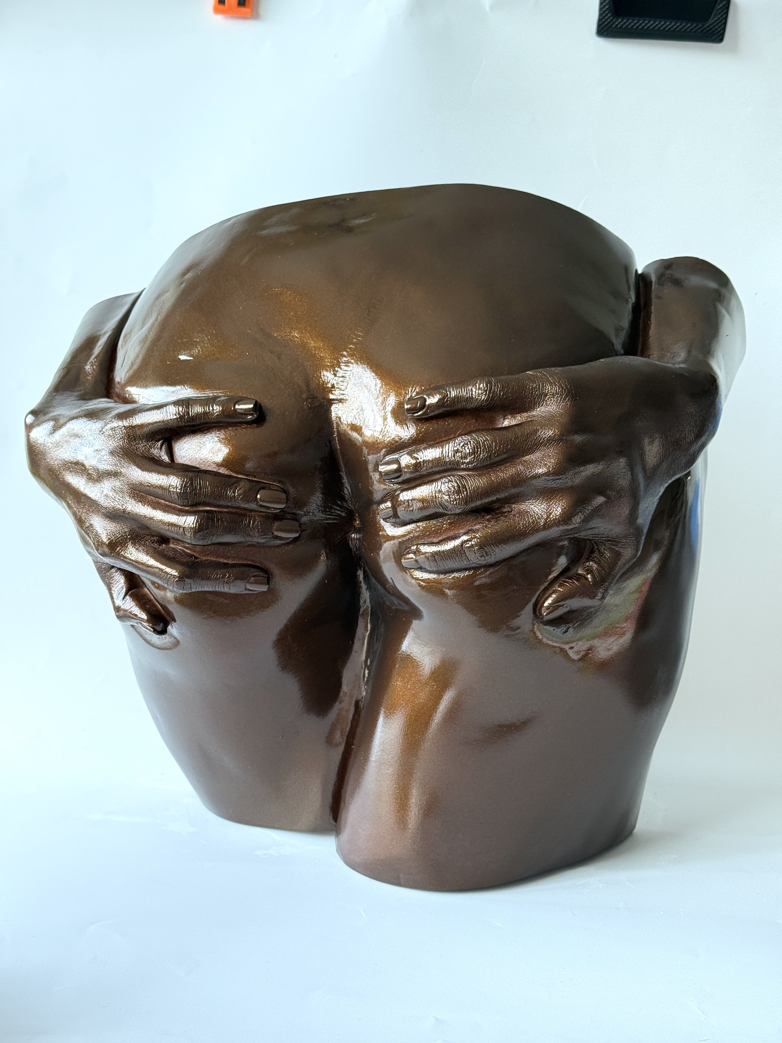 A bronze-colored sculpture of a human head with hands covering the eyes, nose, and mouth, set against a plain white background.