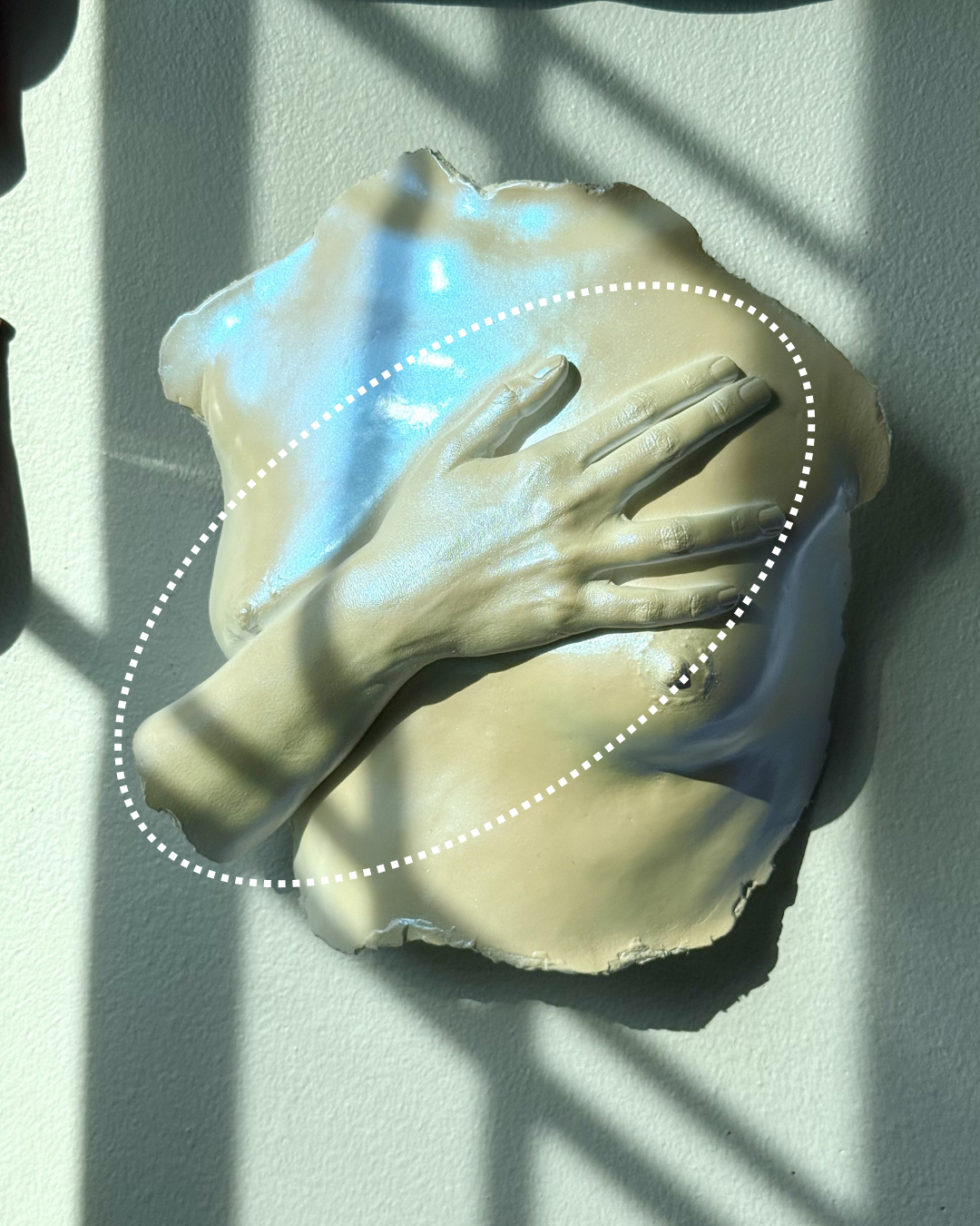 A broken piece of wall with a sculpture of a hand covering a face, placed on a white wall, with shadows cast across the scene.