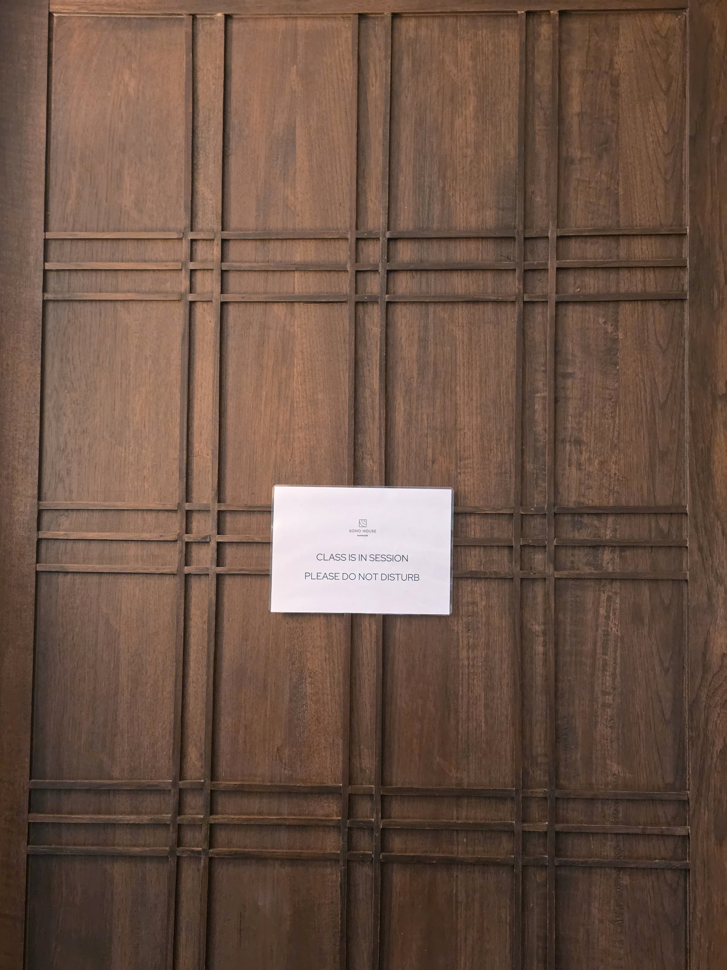 A wooden panelled door at Soho House Bangkok with a white card reading "Class is in session — please do not disturb"