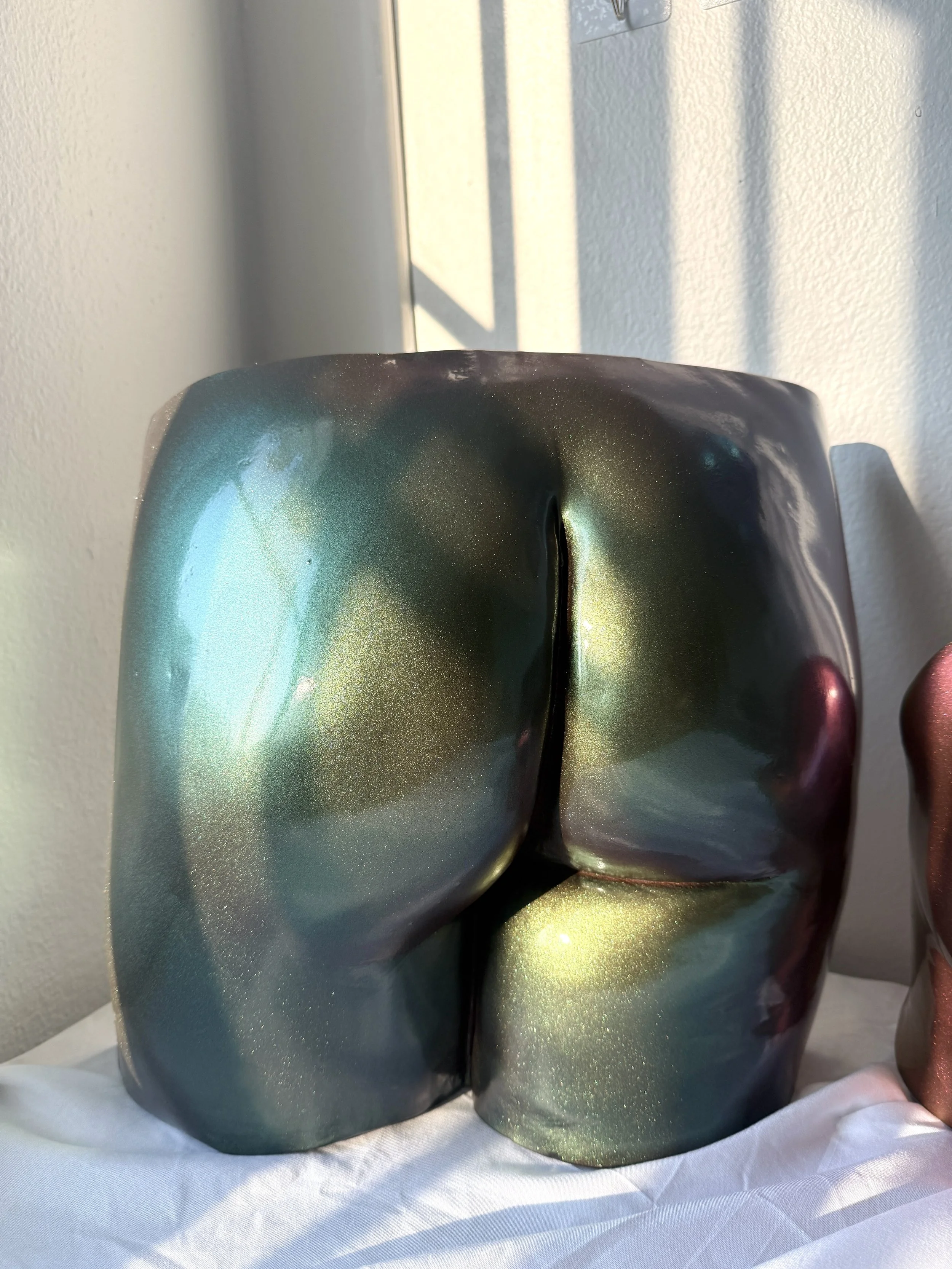 Two metallic, reflective vases with a shiny, iridescent glaze. One appears blue-green, and the other changes colors. They sit on white fabric, with shadows cast by sunlight, against a light-colored wall.