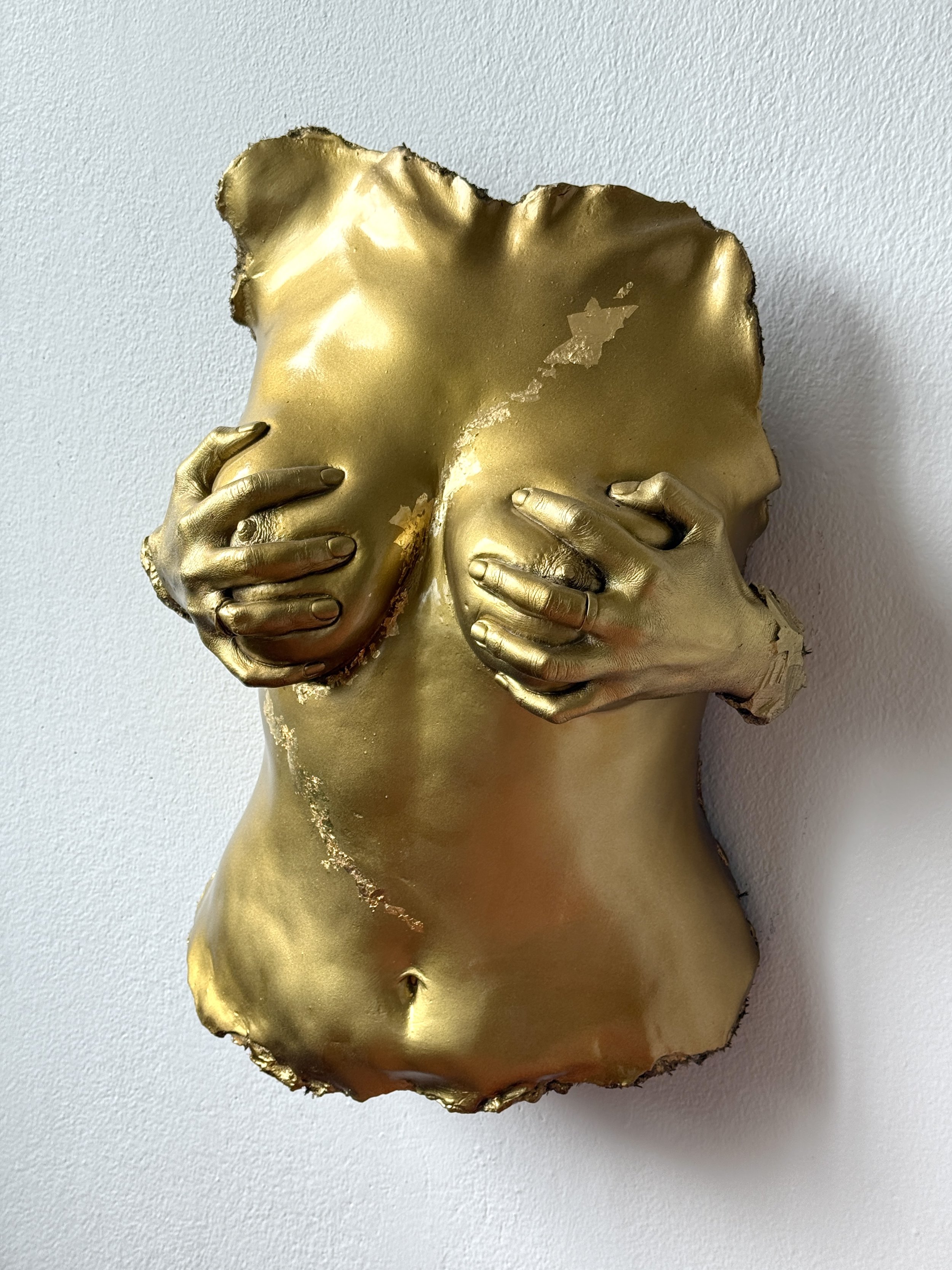 A broken piece of material painted gold, resembling a human torso with hands covering the chest, mounted on a wall.